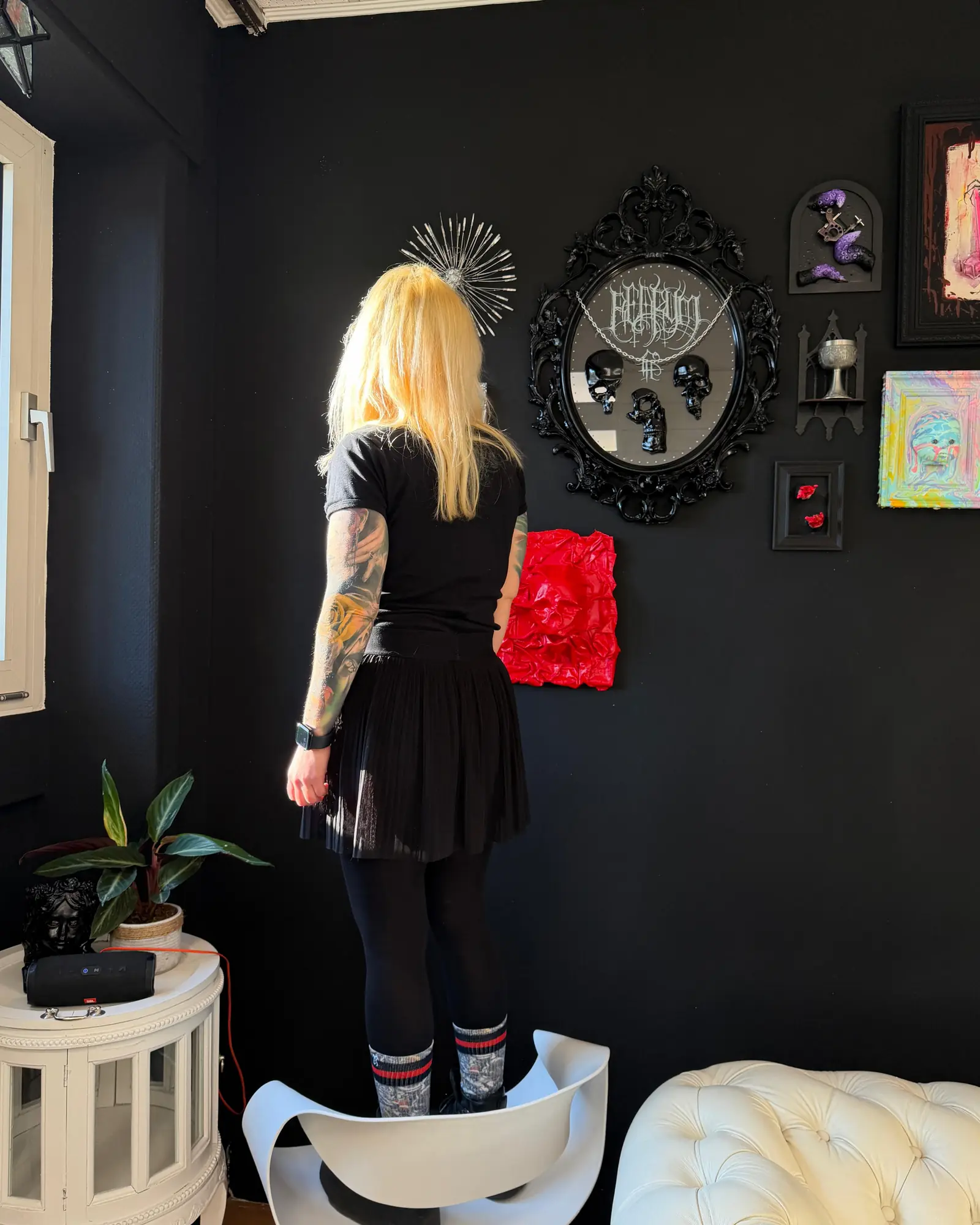 Eine Person steht auf einem Stuhl vor der schwarzen Wand im Studio, daneben ein verzierter Spiegel und gerahmte Kunst.