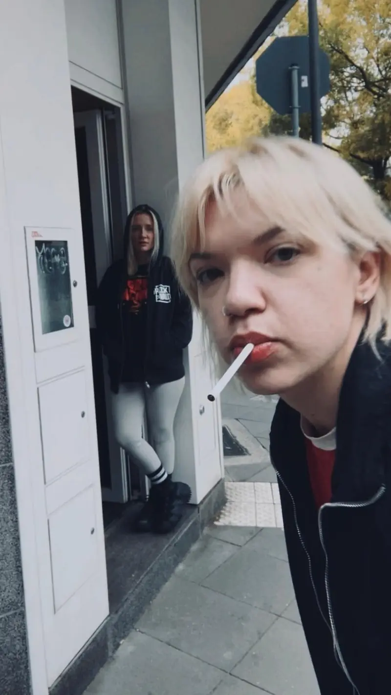 Zwei Personen vor dem Studioeingang an der Straße, eine im Türrahmen, eine im Vordergrund.